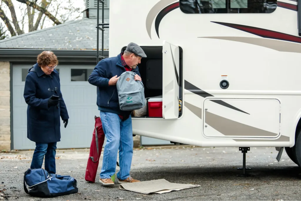 Two people unloading bags from an RV parked outside a house.