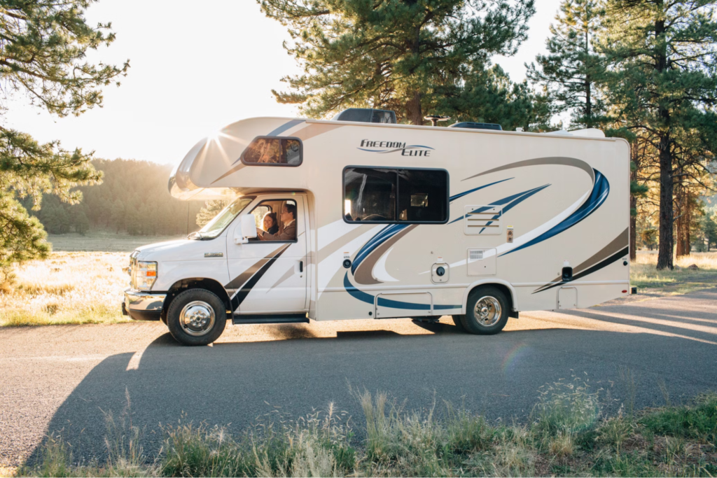 An RV with "Freedom Elite" branding traveling on a road, captured during a sunny day with trees in the background.