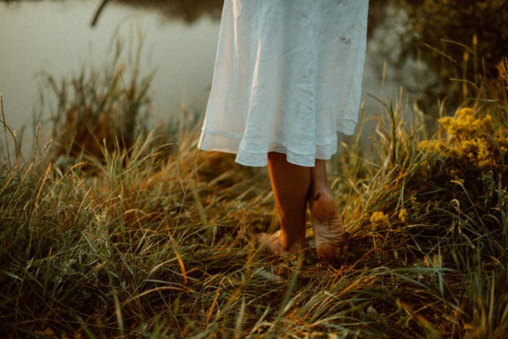 A close-up of a woman standing barefoot on grass, wearing a white dress.