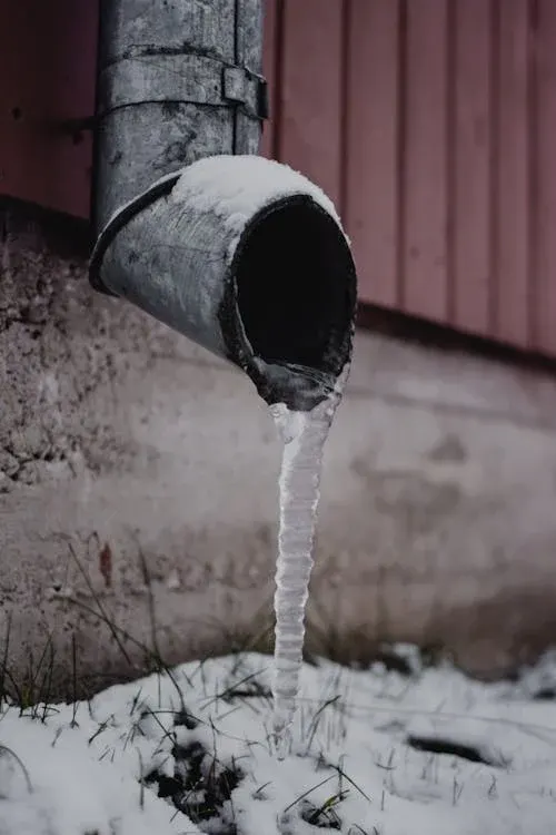 Icicle hanging from the end of a metal drain pipe covered in snow.