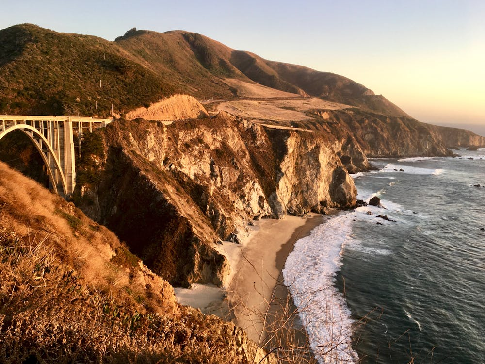 An aerial view of a wooden bridge spanning across a river, surrounded by autumn trees and a sandy shoreline.