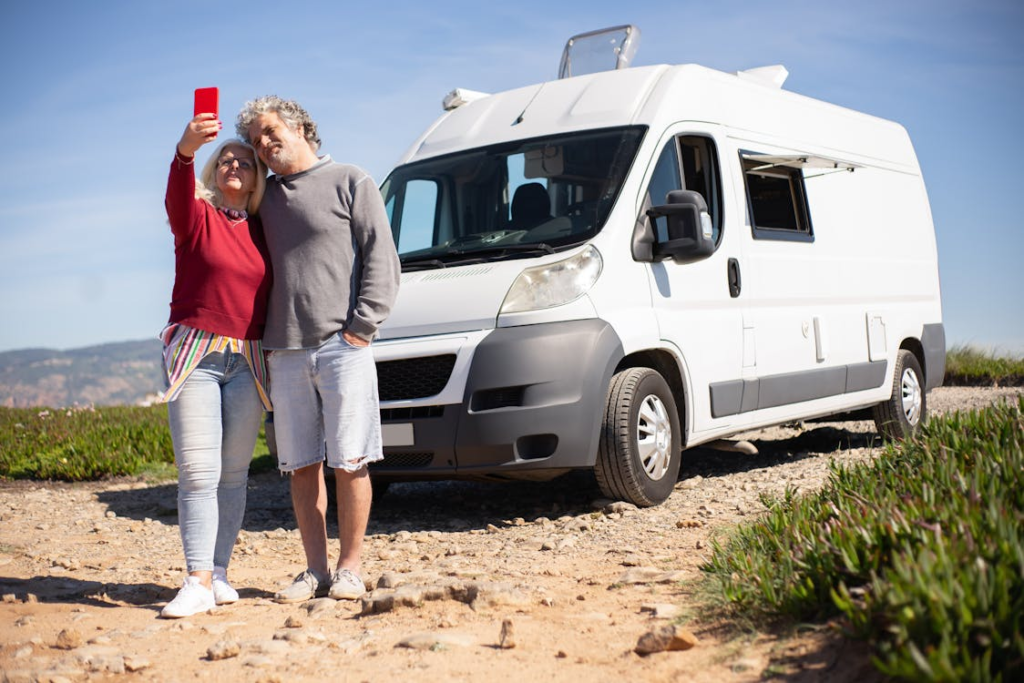  A couple taking a selfie in front of a white camper van parked on a gravel road with a scenic background.