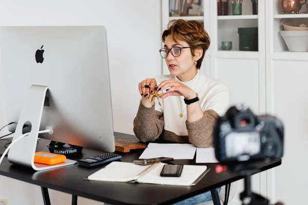 A woman with short hair and glasses sitting at a desk, engaging in a video conference on a large desktop computer, with various office supplies and a camera setup.