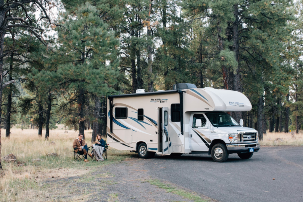 Two people sitting outside of a campervan, which is parked in a forested area.