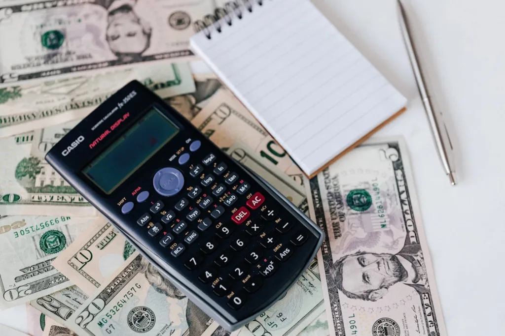 A desk with scattered U.S. dollar bills, a calculator, a notepad, and a pen.