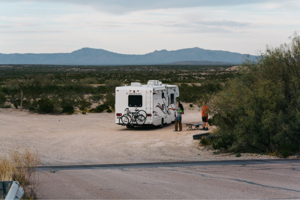 A motorhome parked in a desert area, with two people standing outside it and mountains in the background.