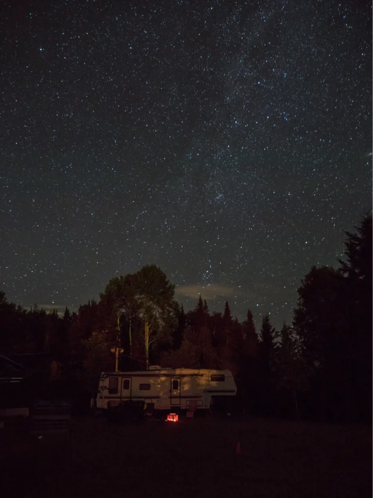 A camper parked in a forest under a starry night sky, with a small campfire glowing underneath.