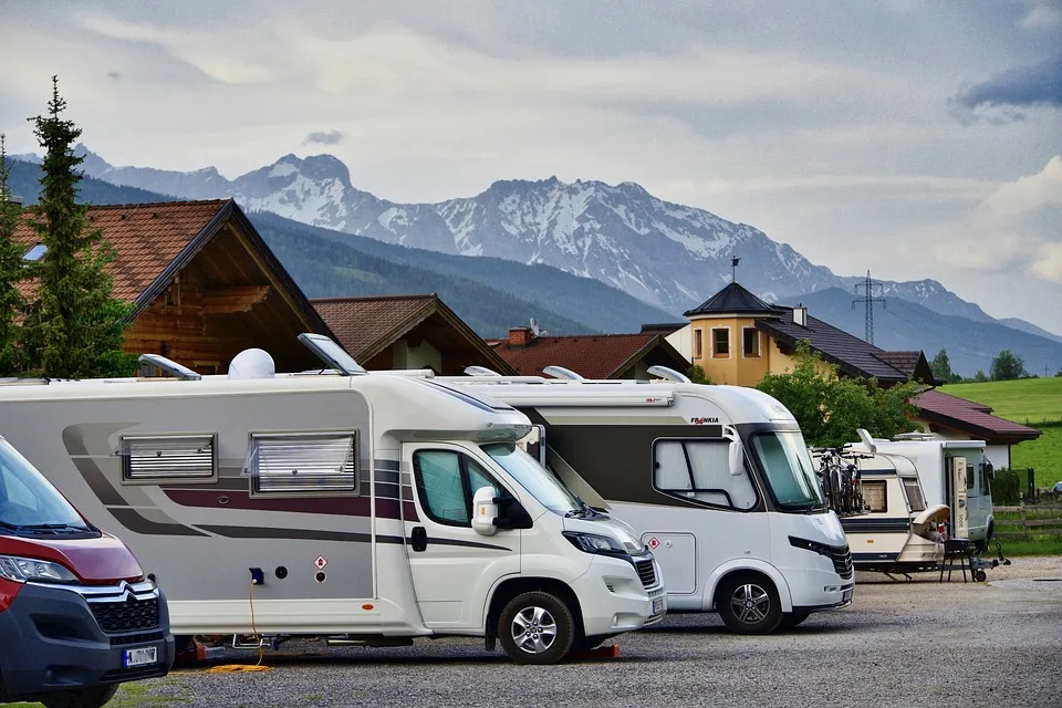 Several motorhomes parked in a campsite with a mountainous backdrop and houses nearby.