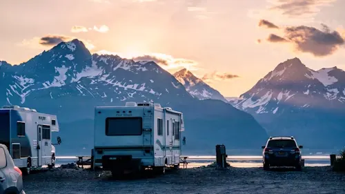 Campervans and a car parked near a body of water with a mountain range in the background during sunset.