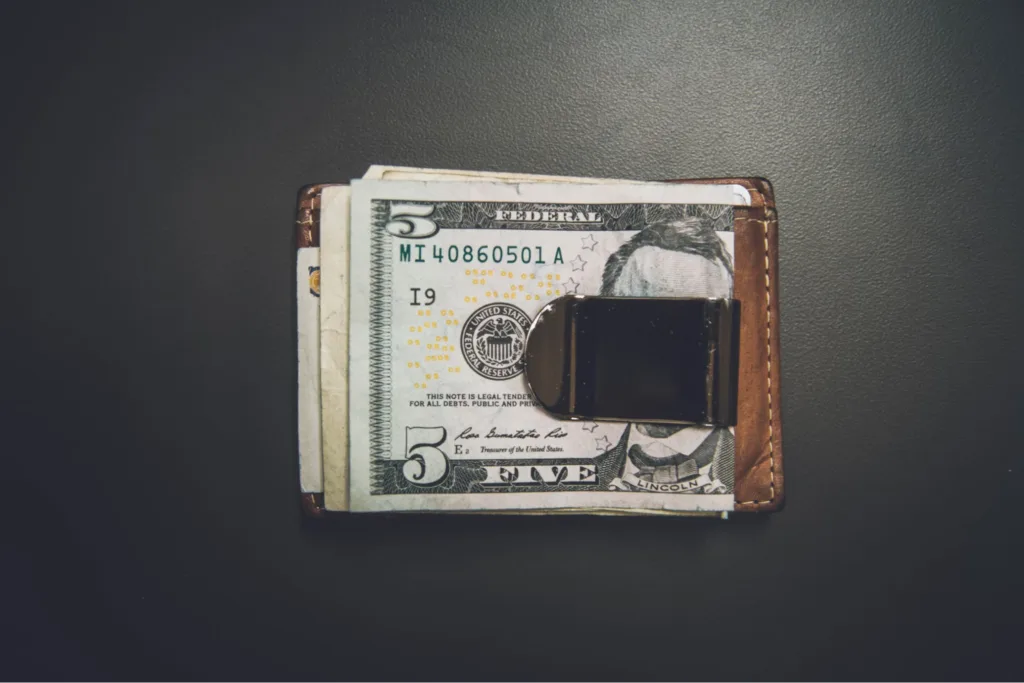 A close-up of a money clip holding several U.S. dollar bills, placed on a dark surface.