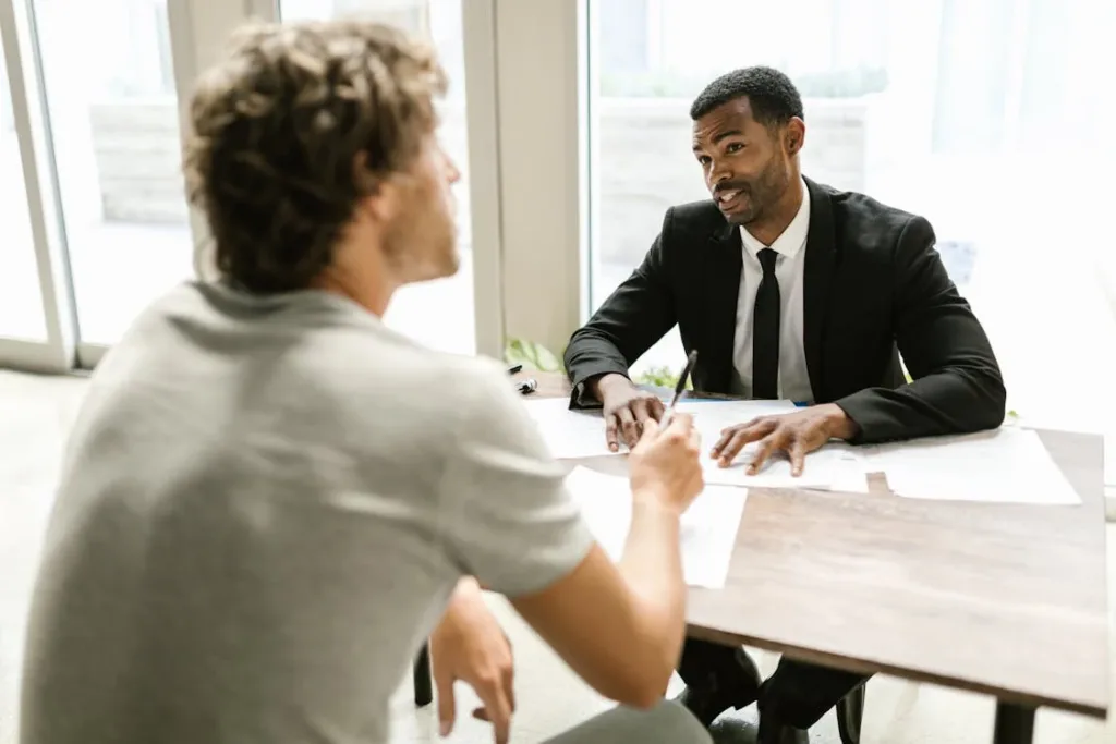 Two people sitting at a table, one in a gray shirt and the other in a black suit, having a discussion.