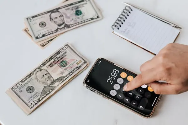 A person using a calculator app on a smartphone to calculate expenses, with stacks of US dollar bills and a notepad in the background.