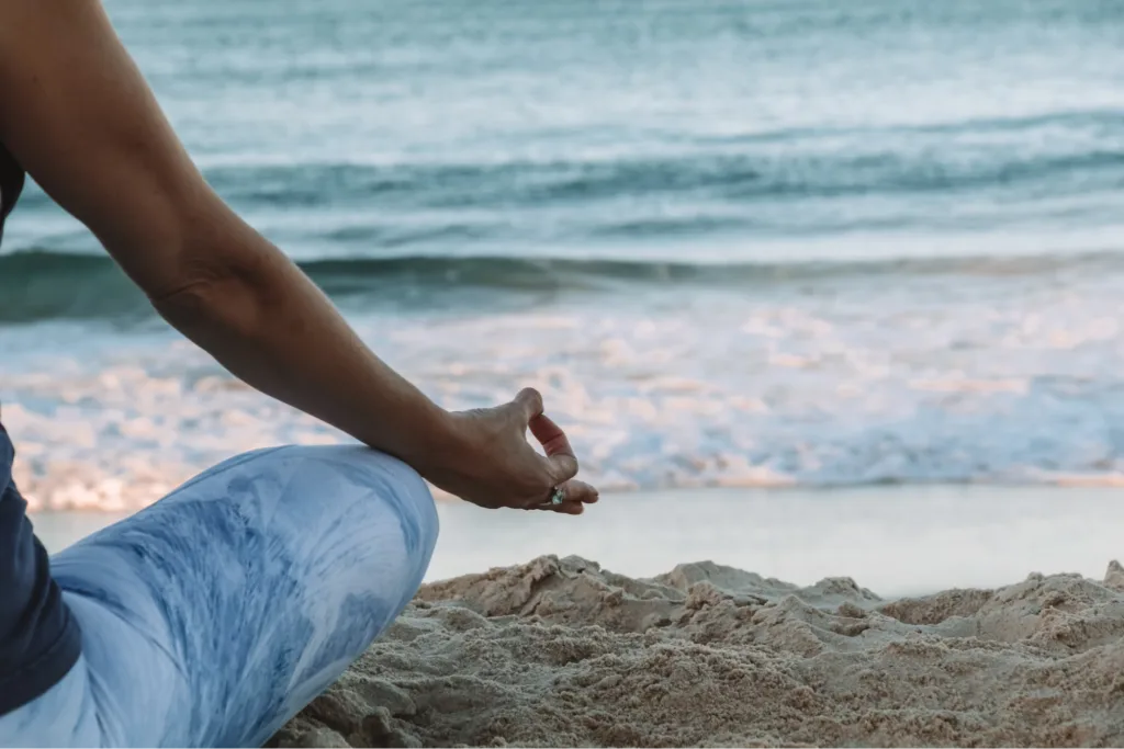 A person practicing yoga or meditation on the beach near the ocean, sitting on the sand with focus on their hands.