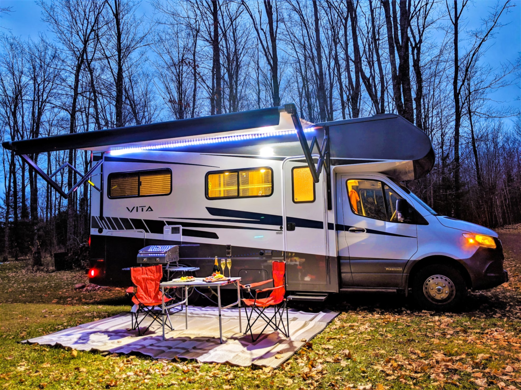 A campervan with "VITA" written on the side is parked in a clearing at dusk. The van has an awning extended with blue led lights, a table set with food and drink, and two camping chairs on a rug.