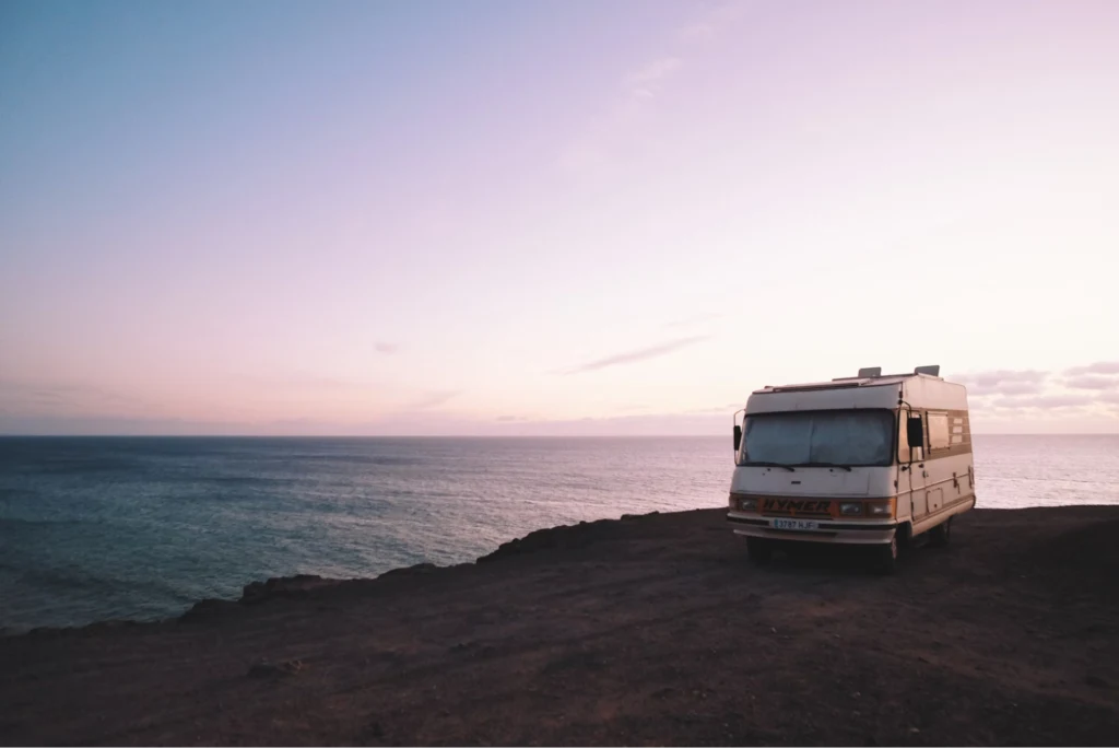 A motorhome parked on a cliff overlooking the ocean at sunrise or sunset, with a clear sky and calm sea.