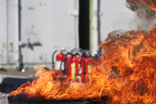 A fire burning intensely with a row of red fire extinguishers in the background.