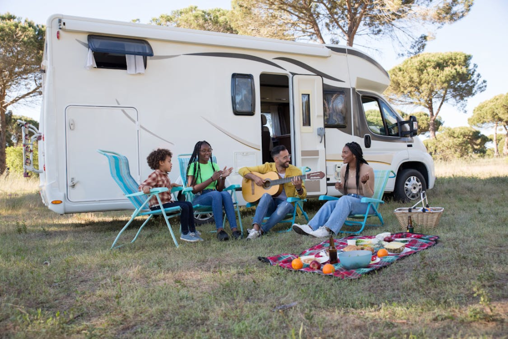 A family sitting on chairs and enjoying a picnic outside a motorhome, with one person playing a guitar, surrounded by trees in a grassy area.