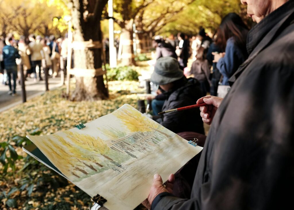 A person painting a scene in a bustling park filled with people and trees in autumn colors.