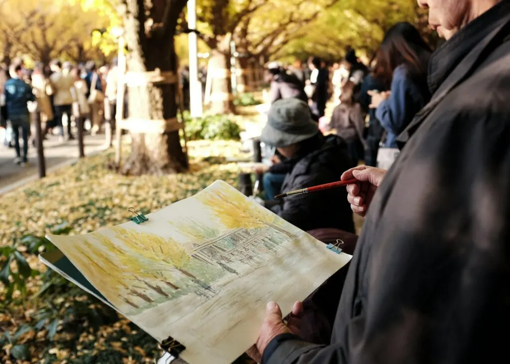 A person painting a scene in a bustling park filled with people and trees in autumn colors.