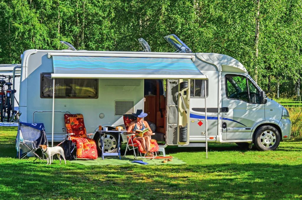 A woman relaxing outside of a camper van parked in a grassy area, with lawn chairs, a table, and a dog nearby.