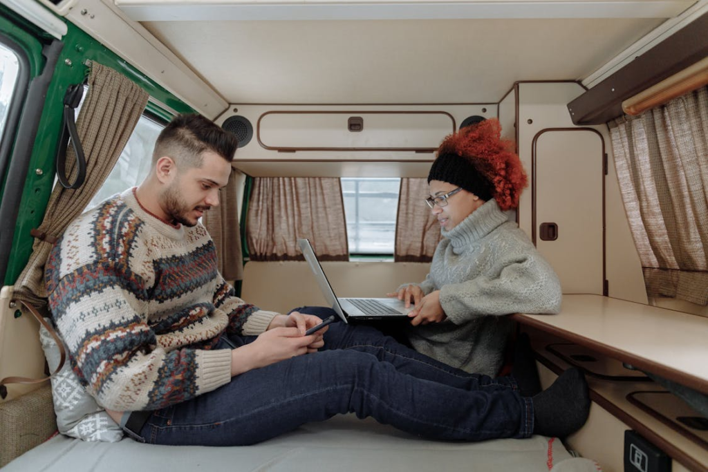 Two people sitting inside a camper van, one using a laptop and the other using a smartphone, both looking cozy in sweaters and surrounded by the van's interior decor.