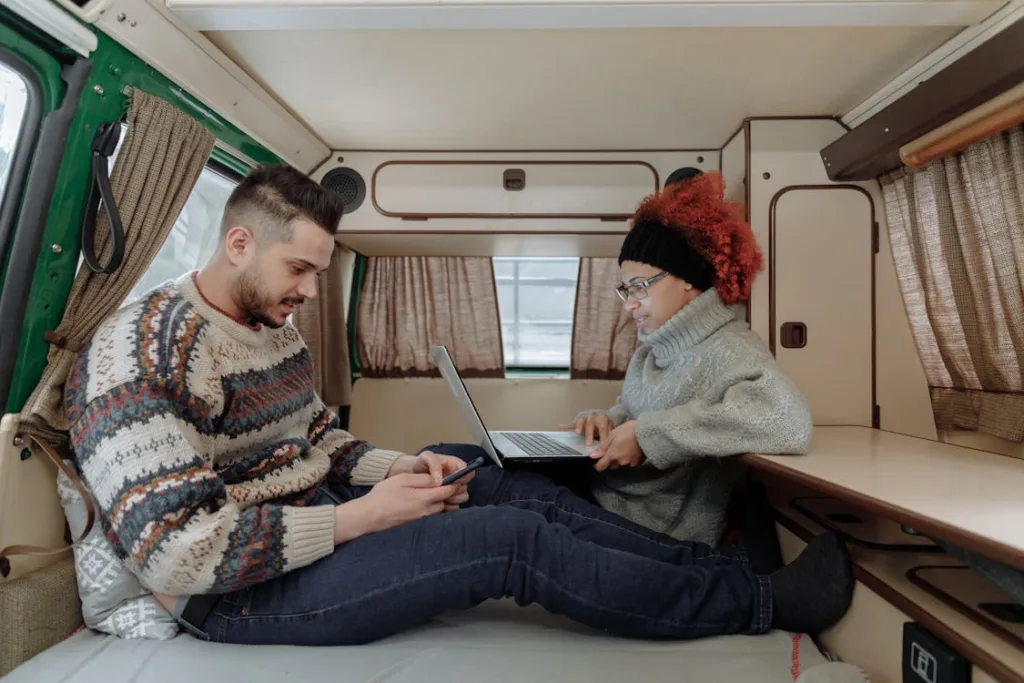 Two people sitting inside a camper van, one using a laptop and the other using a smartphone, both looking cozy in sweaters and surrounded by the van's interior decor.