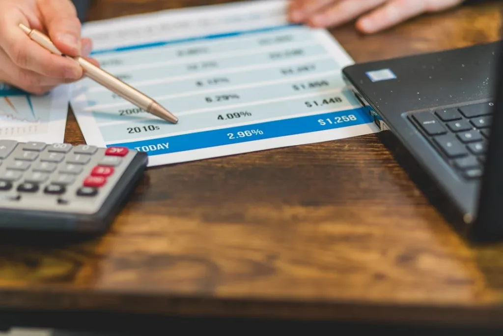 Close-up shot of a person pointing to data on a financial document with a pen, with a calculator and a laptop on a wooden desk.