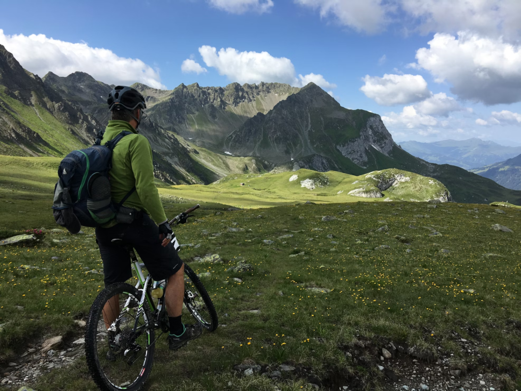 A cyclist standing beside their mountain bike, looking out over a green mountainous landscape on a sunny day.