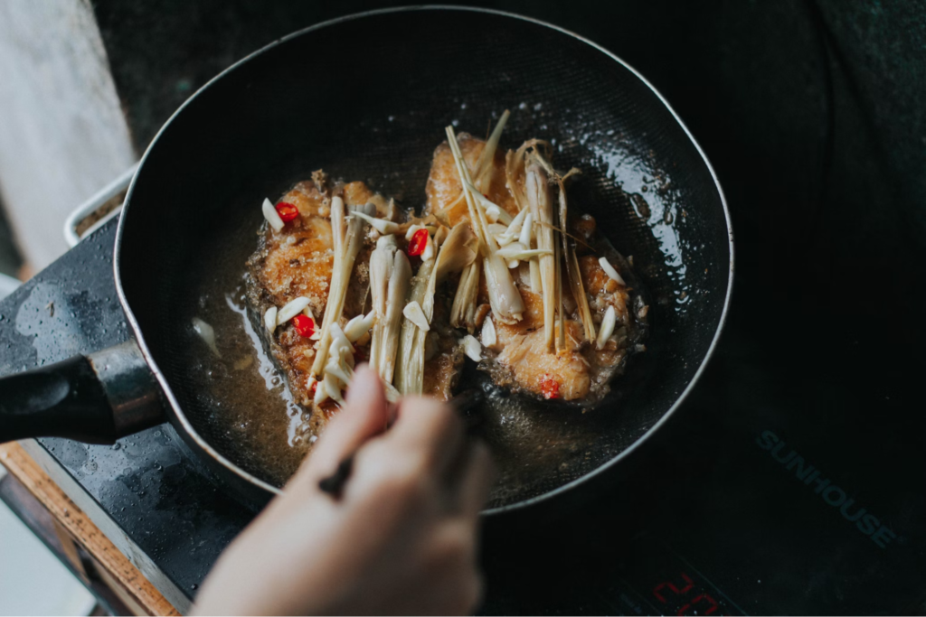 A close-up of salmon being cooked in a frying pan, with various ingredients including garlic, lemongrass, and red chili.