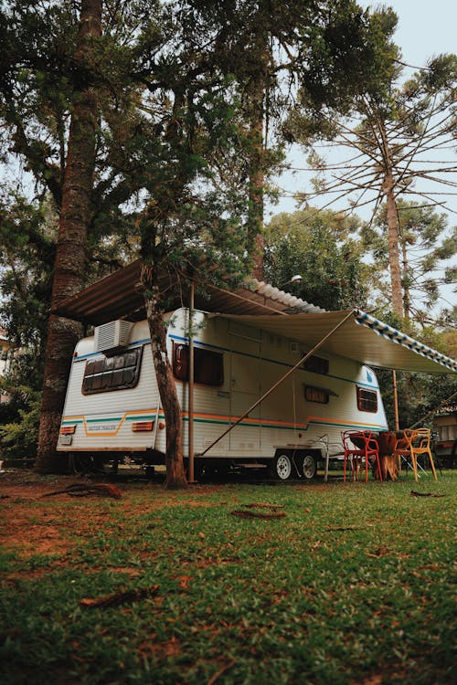 A colorful camper parked on a grassy area among trees, with an extended awning and chairs set up outside.