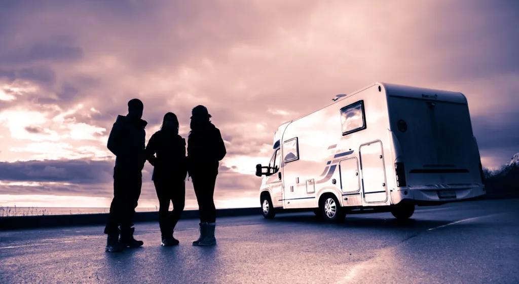 Three silhouetted people standing next to a motorhome at sunset, with dramatic clouds in the background.