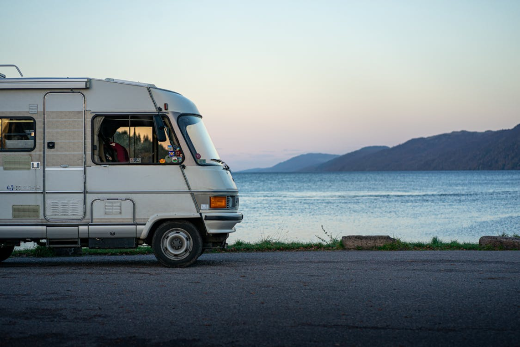 The side view of a motorhome parked by a lake with mountains in the background, under a clear sky.