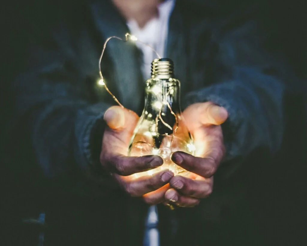 Hands holding a light bulb with tiny lights inside, set against a dark background.