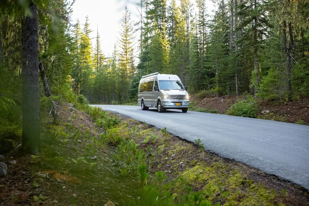 An RV driving on a scenic forest road, surrounded by trees with sunlight filtering through.