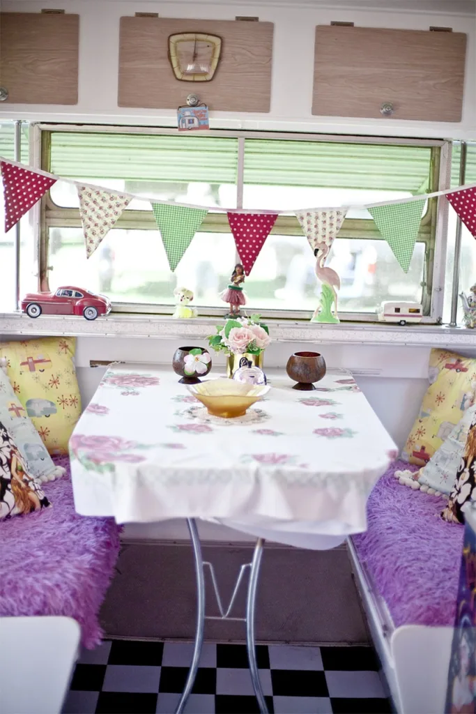 A cheerfully decorated camper van interior featuring a dining area with a flowery tablecloth, colorful cushions, and decorative bunting hanging above.