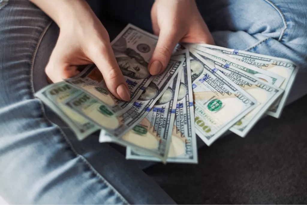 A person holding a fan of hundred-dollar bills over their lap, wearing blue jeans.