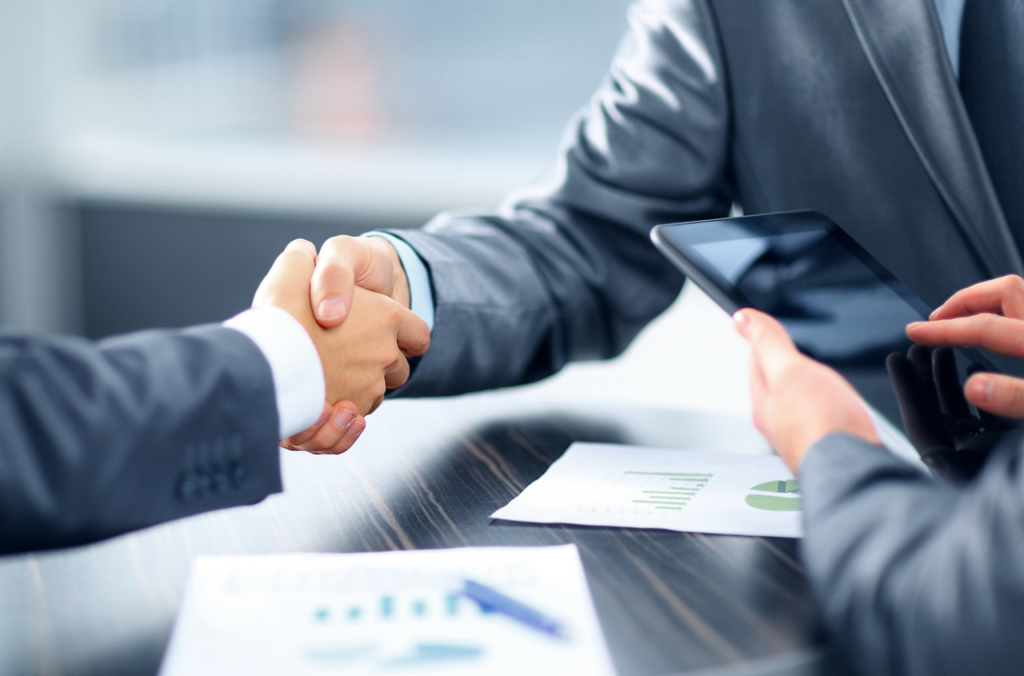 Two people in business attire shaking hands, with one holding a tablet and financial documents on the table.