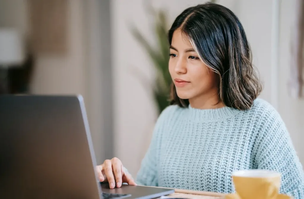 A woman with shoulder-length hair wearing a light blue sweater, concentrating while using a laptop, with a cup beside her.