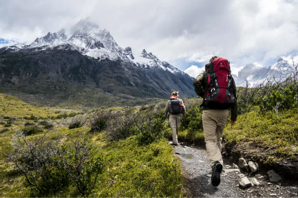 Two hikers with backpacks walking along a trail through a valley, with snow-capped mountains in the background.