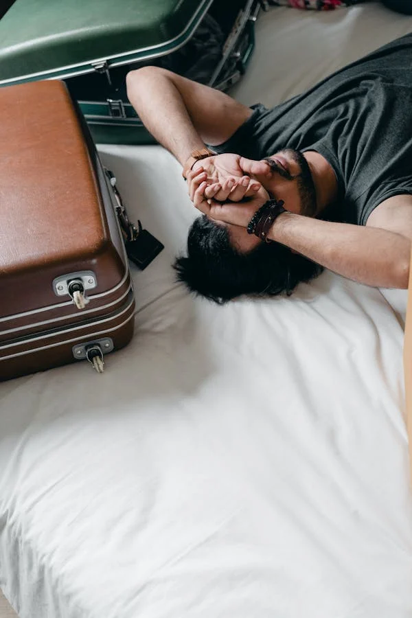 An overhead view of a person lying on a bed with their eyes closed, surrounded by suitcases