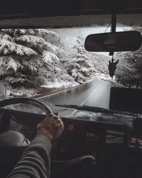 View from inside a vehicle showing a driver’s hands on the steering wheel, driving through a scenic snowy landscape.