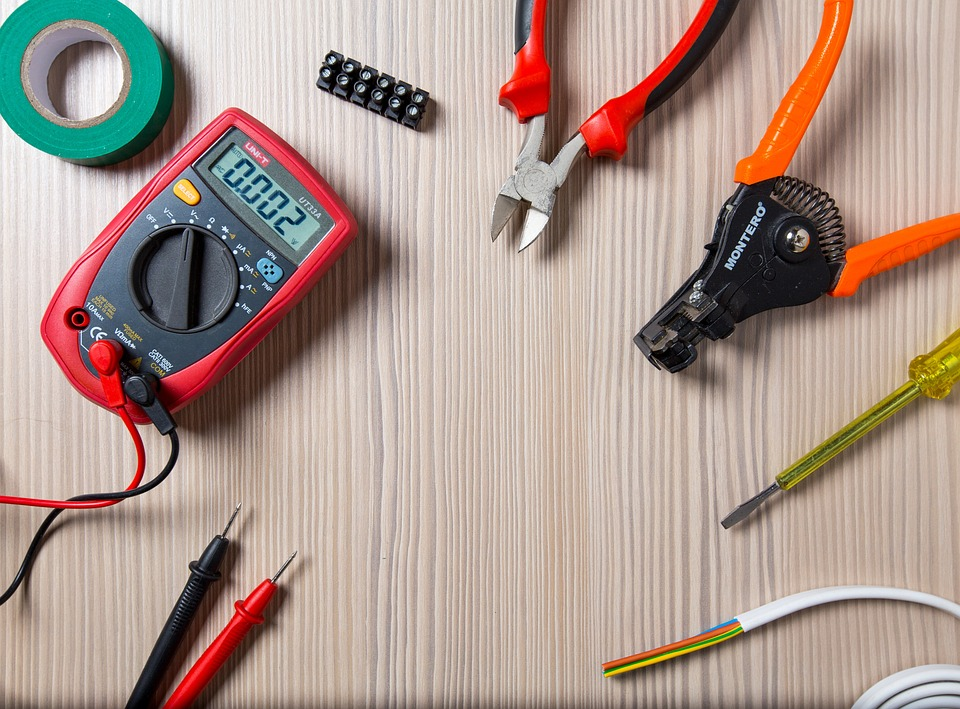 A set of tools laid out on a wooden surface, including a multimeter, wire strippers, pliers, a screwdriver, electrical tape, a wire connector block, and a length of wire.