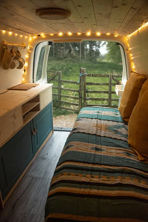 Cozy interior of a campervan with a bed, string lights, and a small cabinet, looking out onto a green yard with a fence.