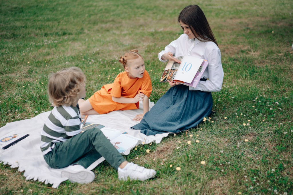 Three children sitting on a blanket in the grass while a young woman reads to them from a book.