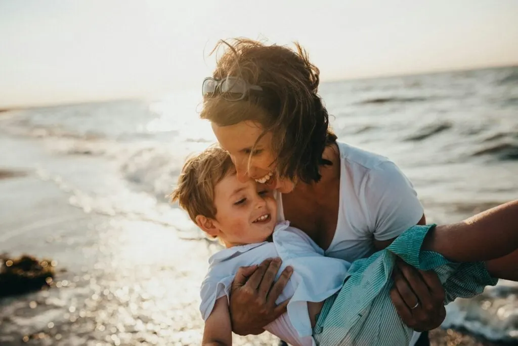 A smiling woman holding a child by the seaside, with waves crashing in the background.