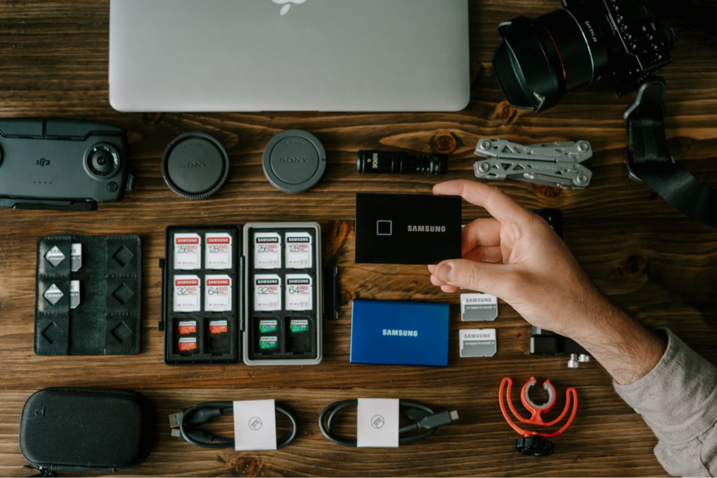 An overhead view of various photography and tech gear, including SD cards, camera lenses, hard drives, and cables, neatly arranged on a wooden table.