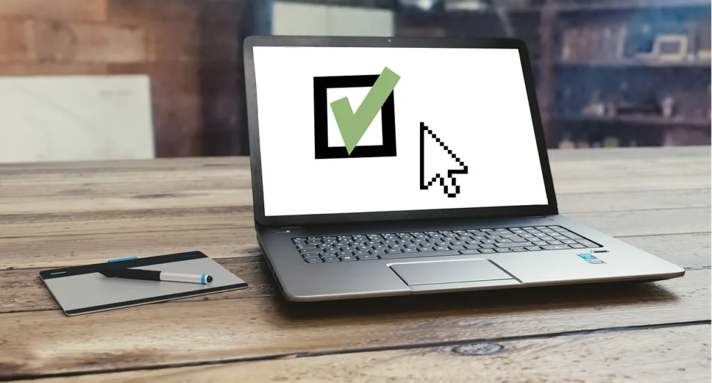 A laptop on a wooden desk showing a screen with a large green checkmark inside a checkbox, next to a digital tablet stylus pad.