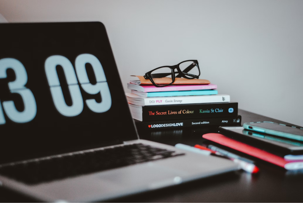 A close-up of a laptop screen showing the time, with a stack of books and a pair of glasses on a desk.