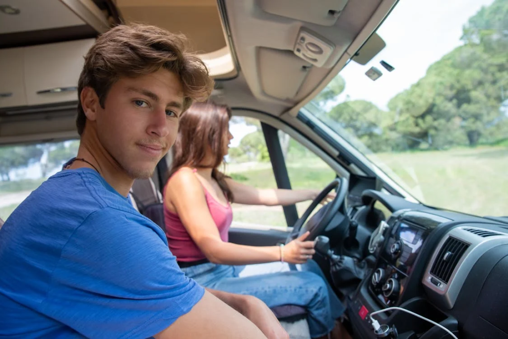 Two people inside an RV; woman is driving while the man looks at the camera.