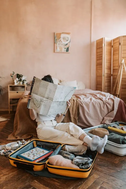 A person sitting cross-legged on the floor with an open suitcase, reading a large map in a cozy bedroom.